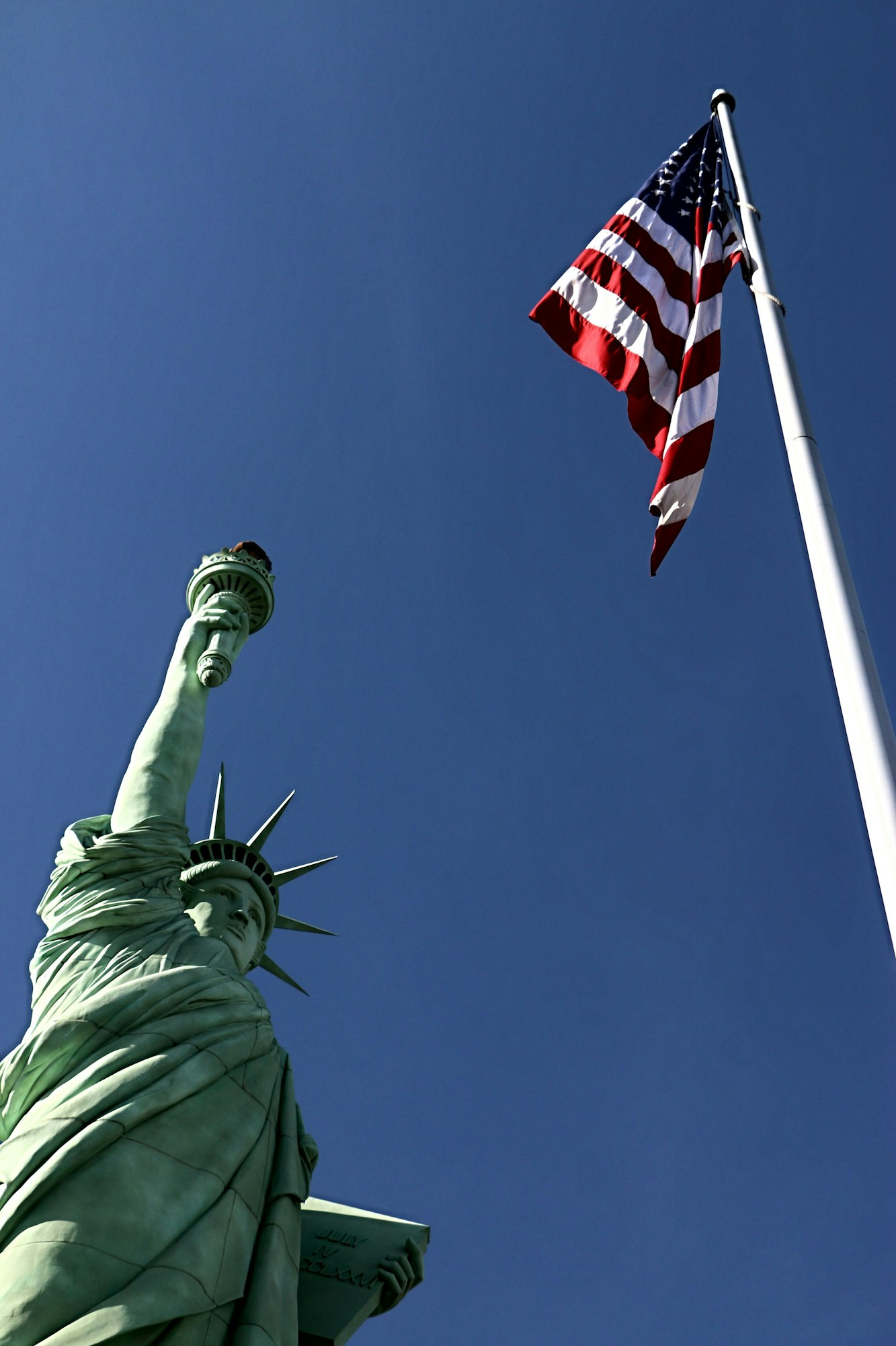 Vertical image of the Statue of Liberty and an American flag against a clear blue sky.